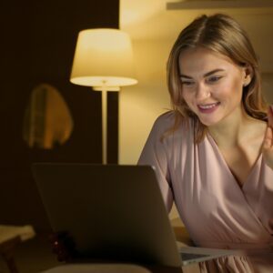 Woman smiling and waving during a video call, using a laptop in a cozy indoor setting.